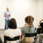 Man presenting with tablet to a small seated audience in a modern indoor setting.