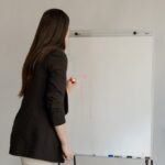 Female employee in black blazer writing on whiteboard during office meeting.
